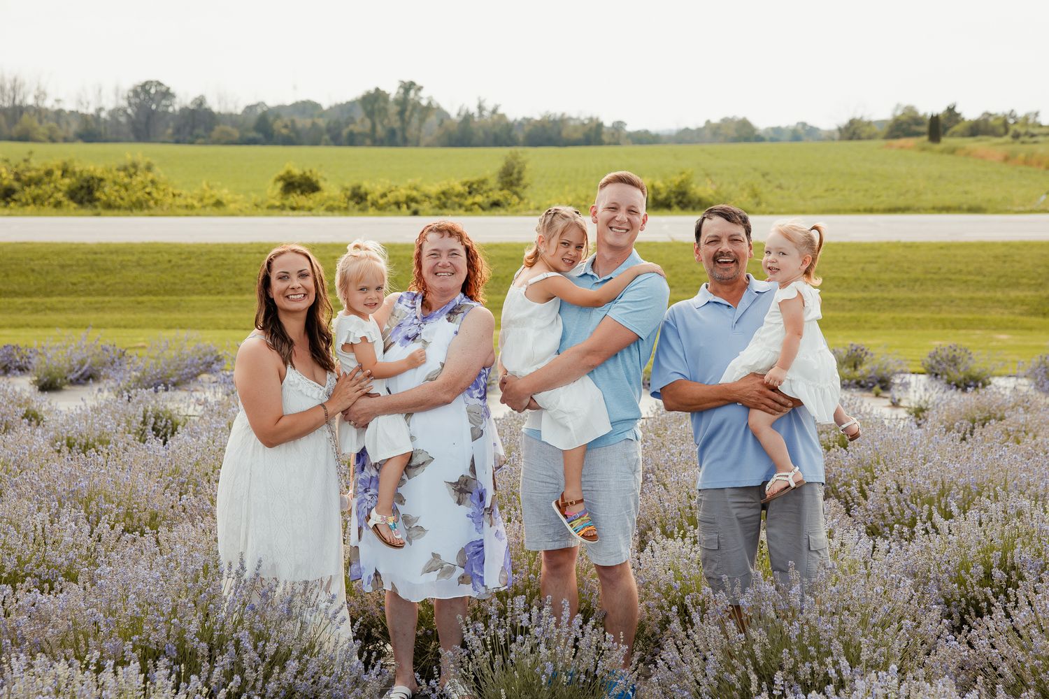 Family portrait of seven people smiling in a lavender field with green farmland in the background.