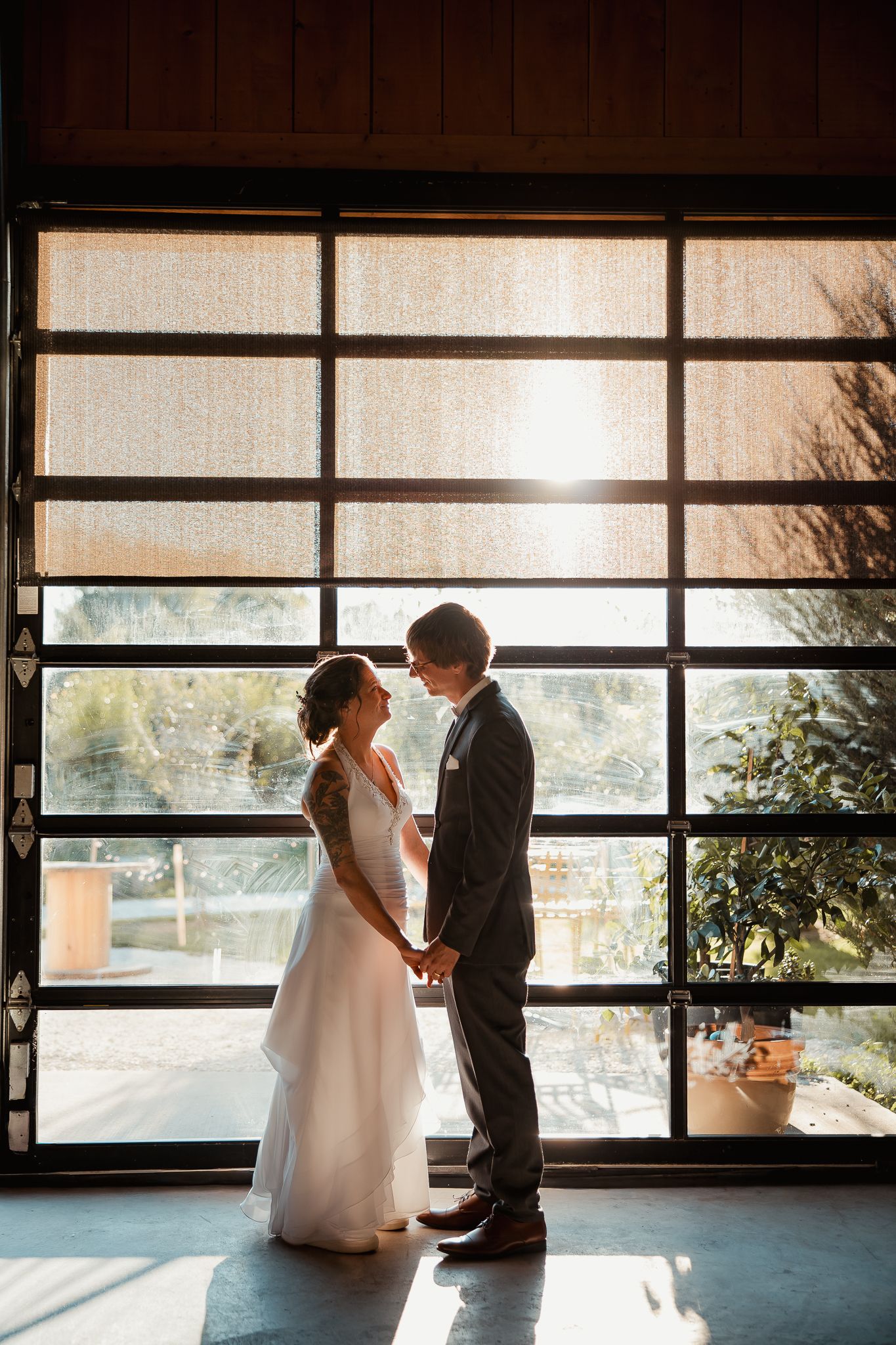 A romantic silhouette of a couple in wedding attire stands before large windows in a modern venue with natural light.