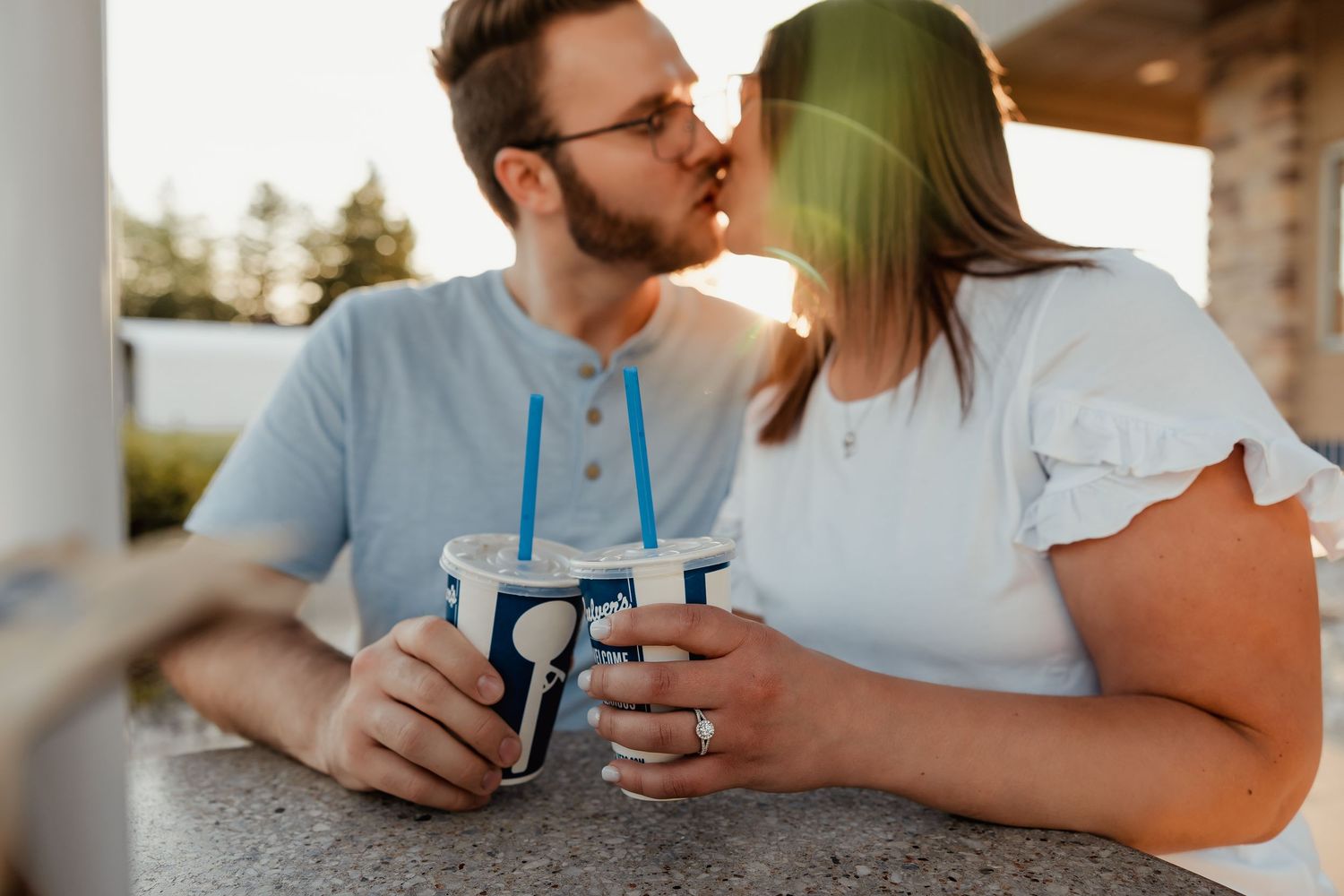 Couple sharing a kiss while holding matching blue striped drinks at an outdoor cafe during golden hour.