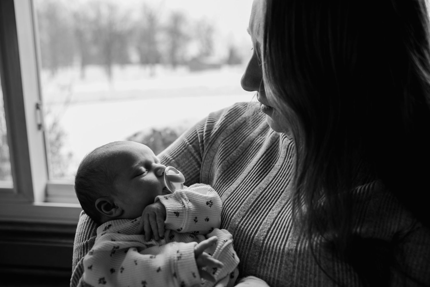 A tender black and white moment between parent and newborn baby by a window in soft natural light.