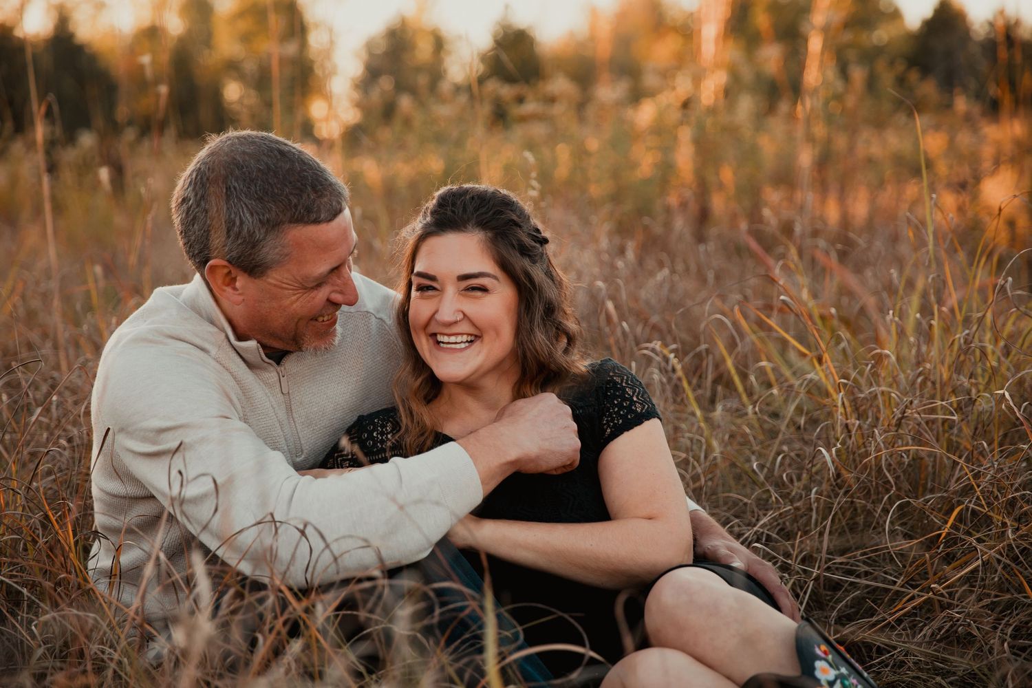 A couple laughs together while sitting in a golden field during sunset. couples engagement