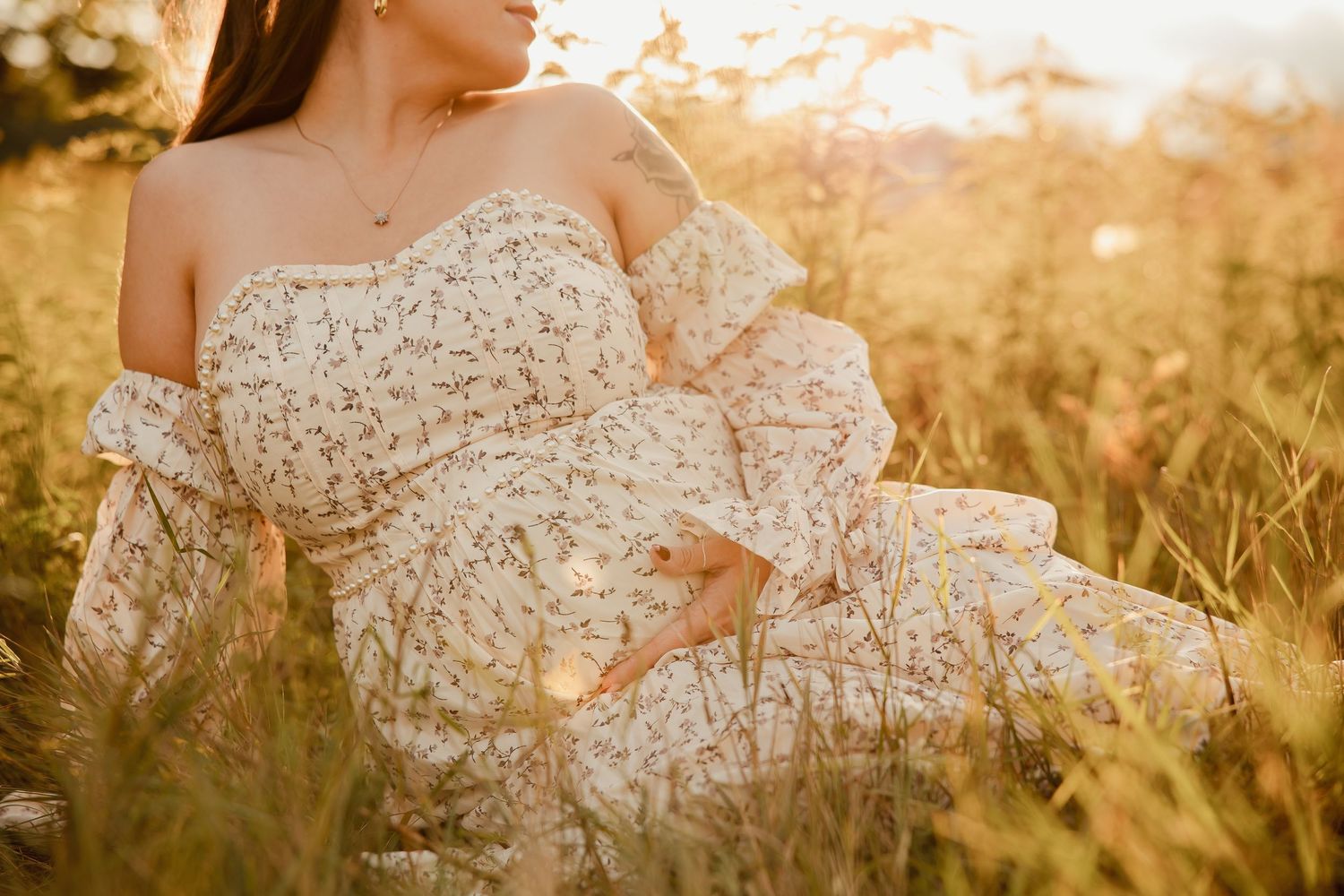 Woman in floral off-shoulder dress sitting in golden sunlit meadow grass during sunset.