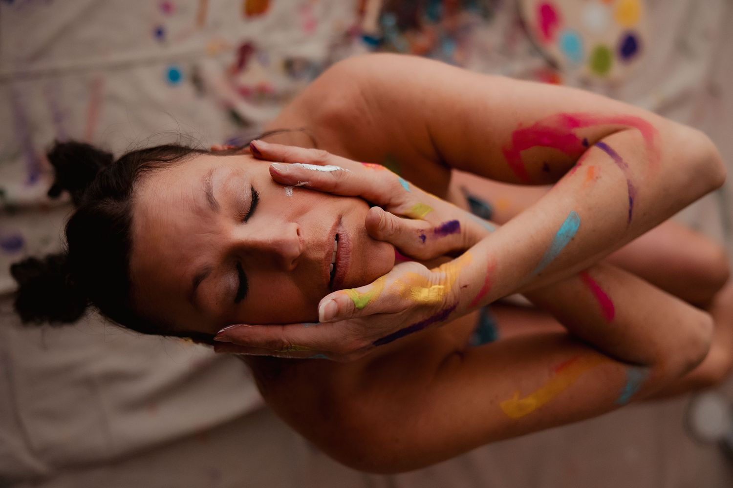Woman with colorful paint on her hands and body lying down with eyes closed in artistic body paint photography.