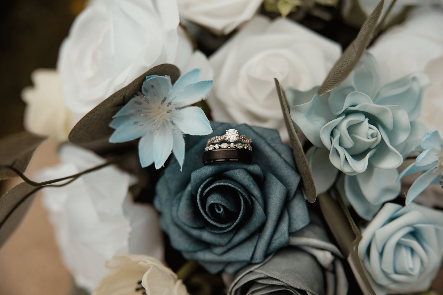 An engagement ring nestled between blue and white roses in a wedding bouquet shows subtle details and sparkle.