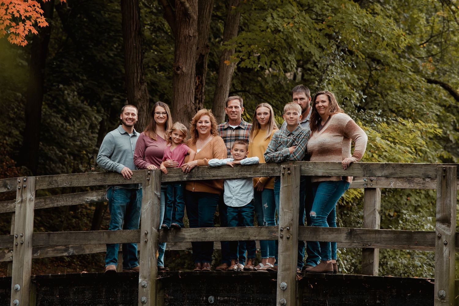 Large family group portrait on a wooden bridge surrounded by autumn foliage, wearing coordinated casual outfits.