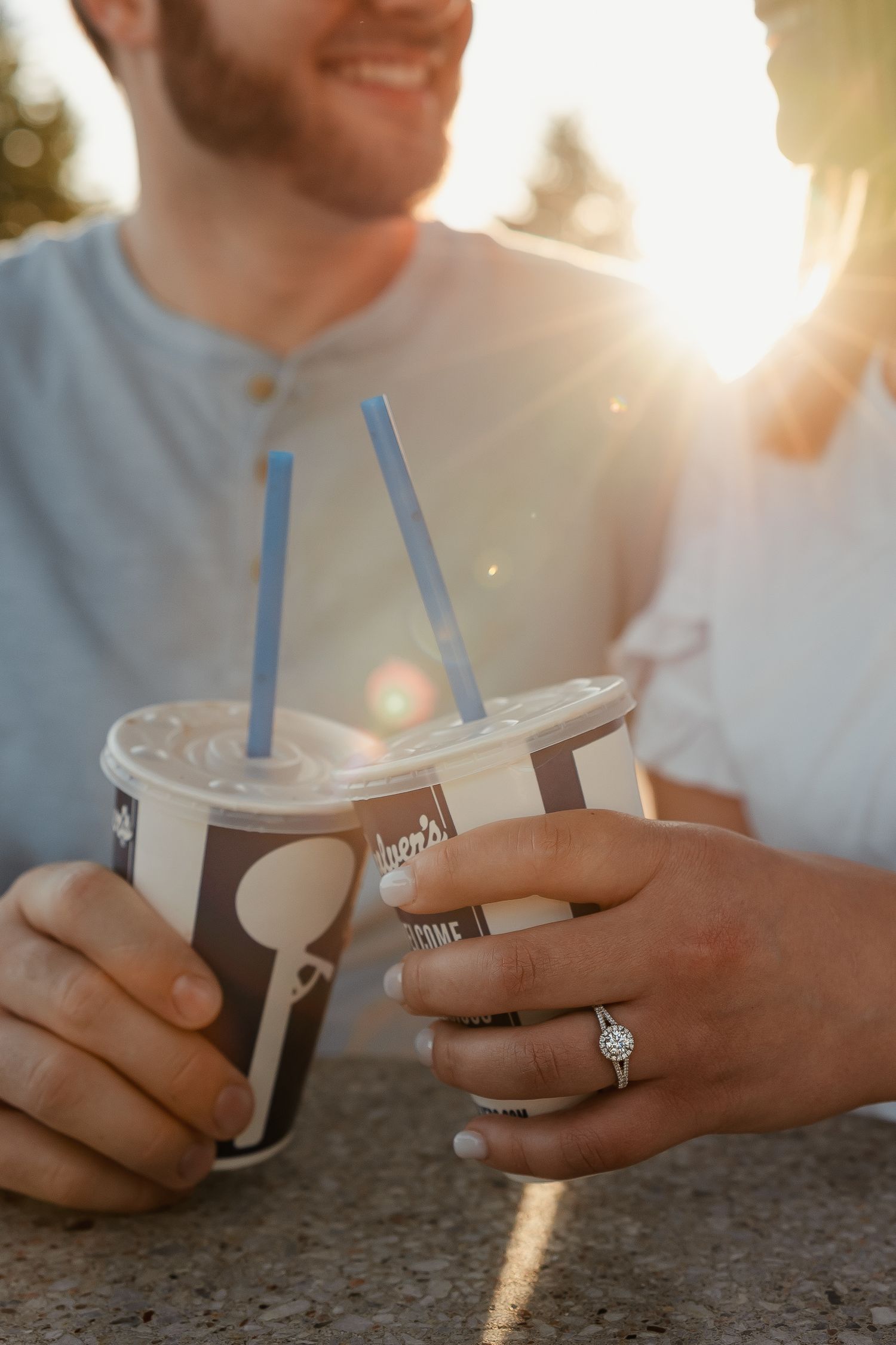 Engaged couple holding Culvers frozen custard cups with diamond ring visible in golden sunlight.