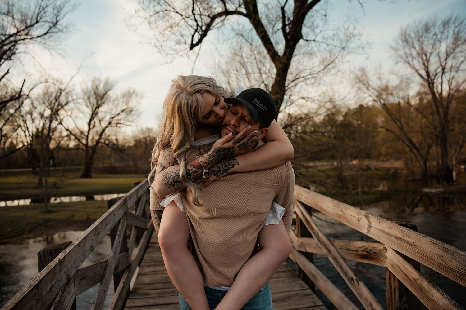 A couple shares a romantic piggyback embrace on a wooden bridge surrounded by autumn trees at sunset.
