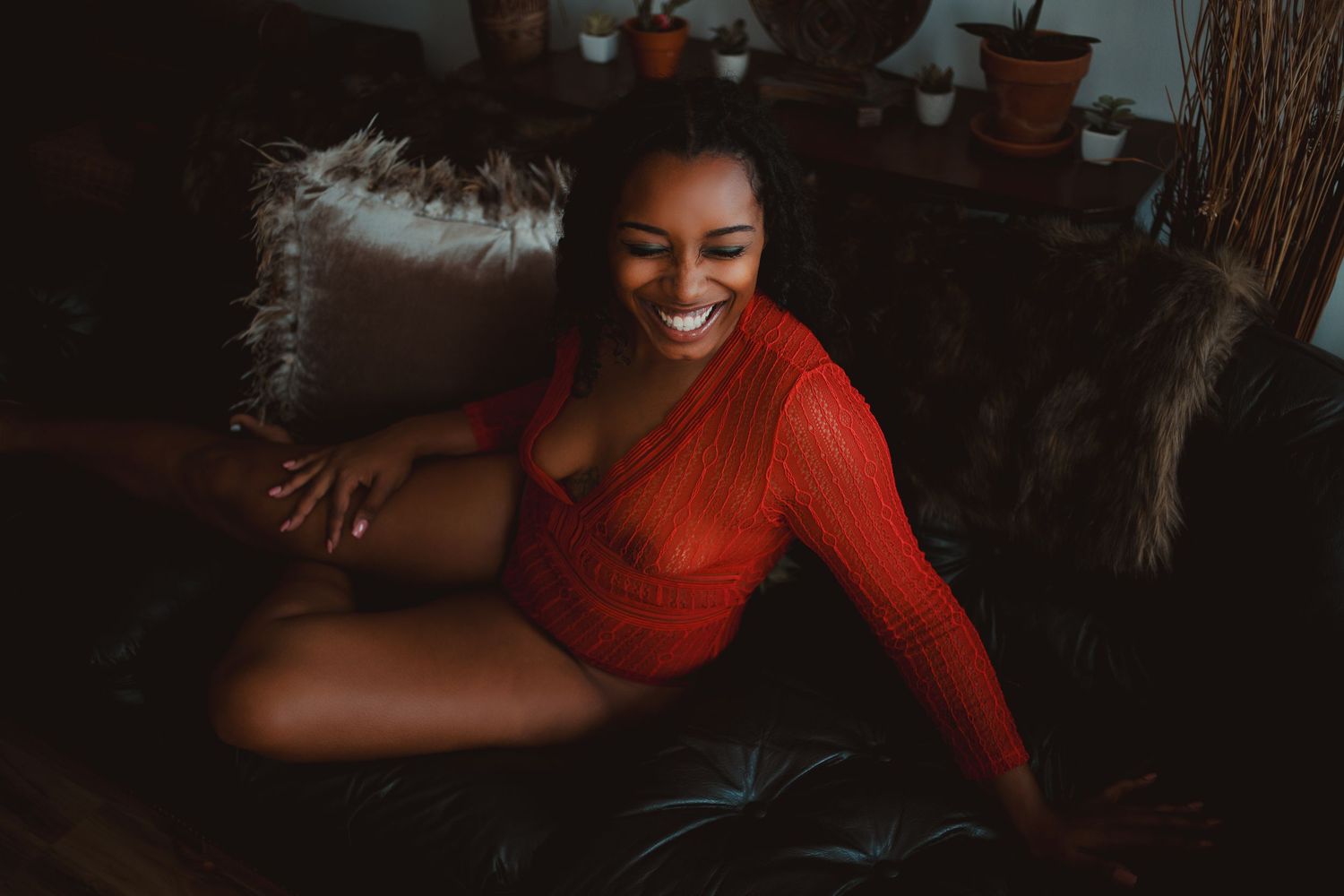 A person in a red sweater sits comfortably on a dark couch with a warm smile. boudoir