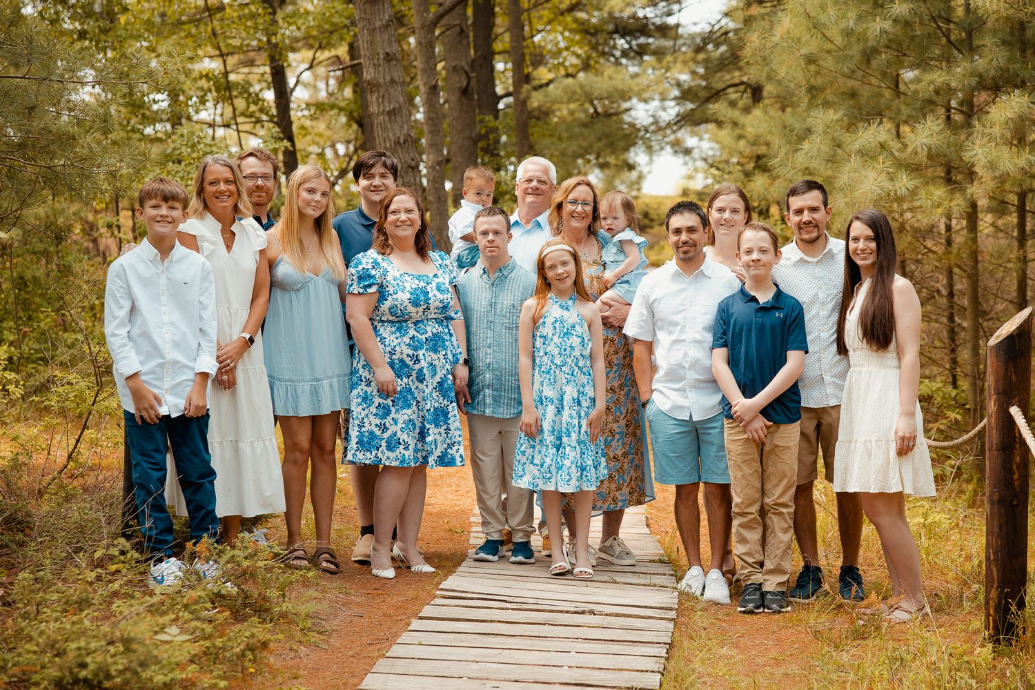 Large family group portrait on a wooden bridge path surrounded by pine trees, dressed in coordinating blue and white outfits.