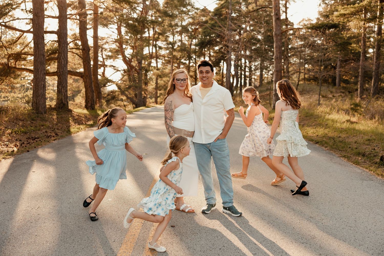 Family of six poses on a sunlit forest road, parents surrounded by four daughters in pastel dresses during golden hour.