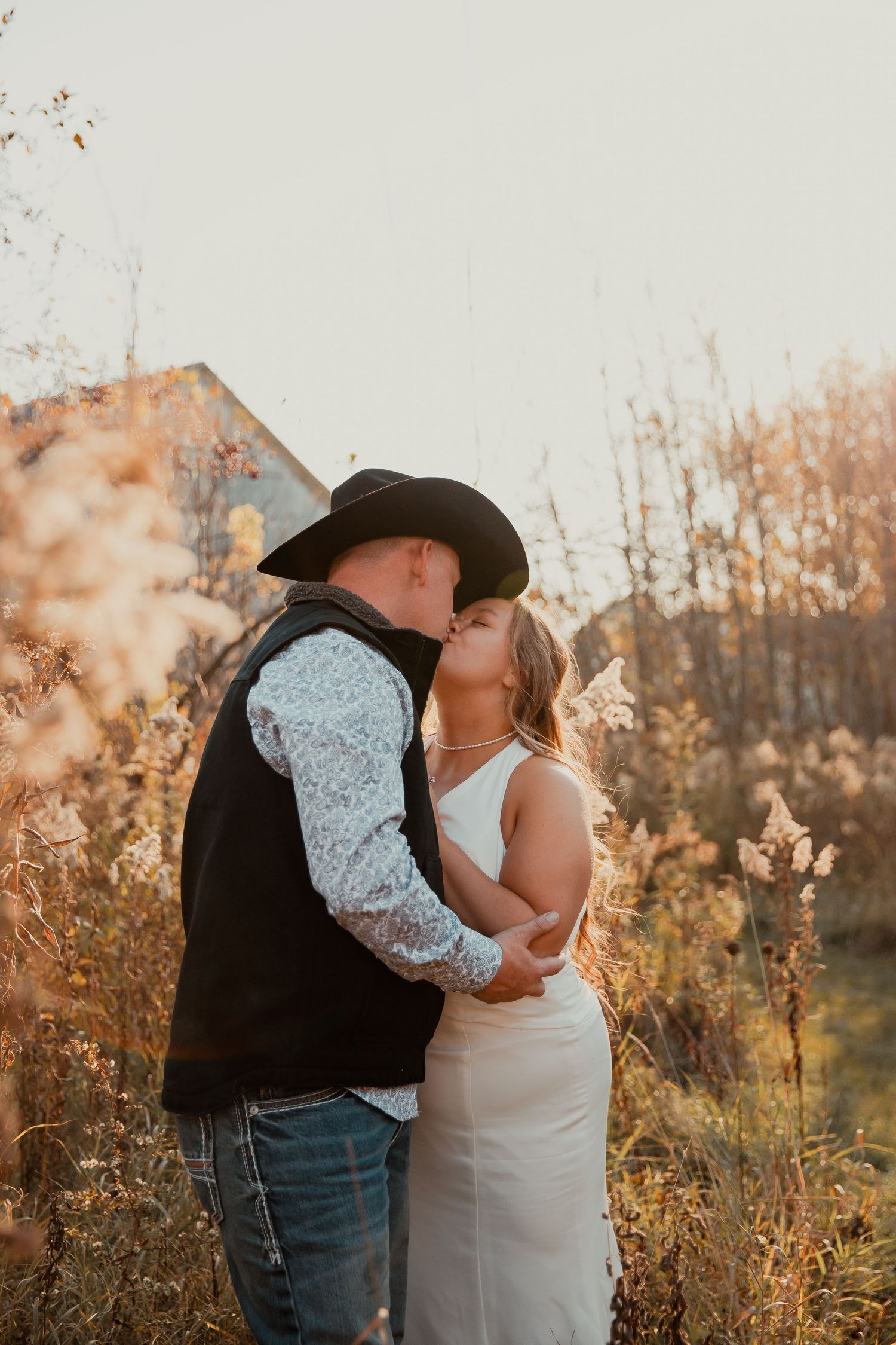Couple in western wedding attire embracing in a field of tall grass during sunset. wedding