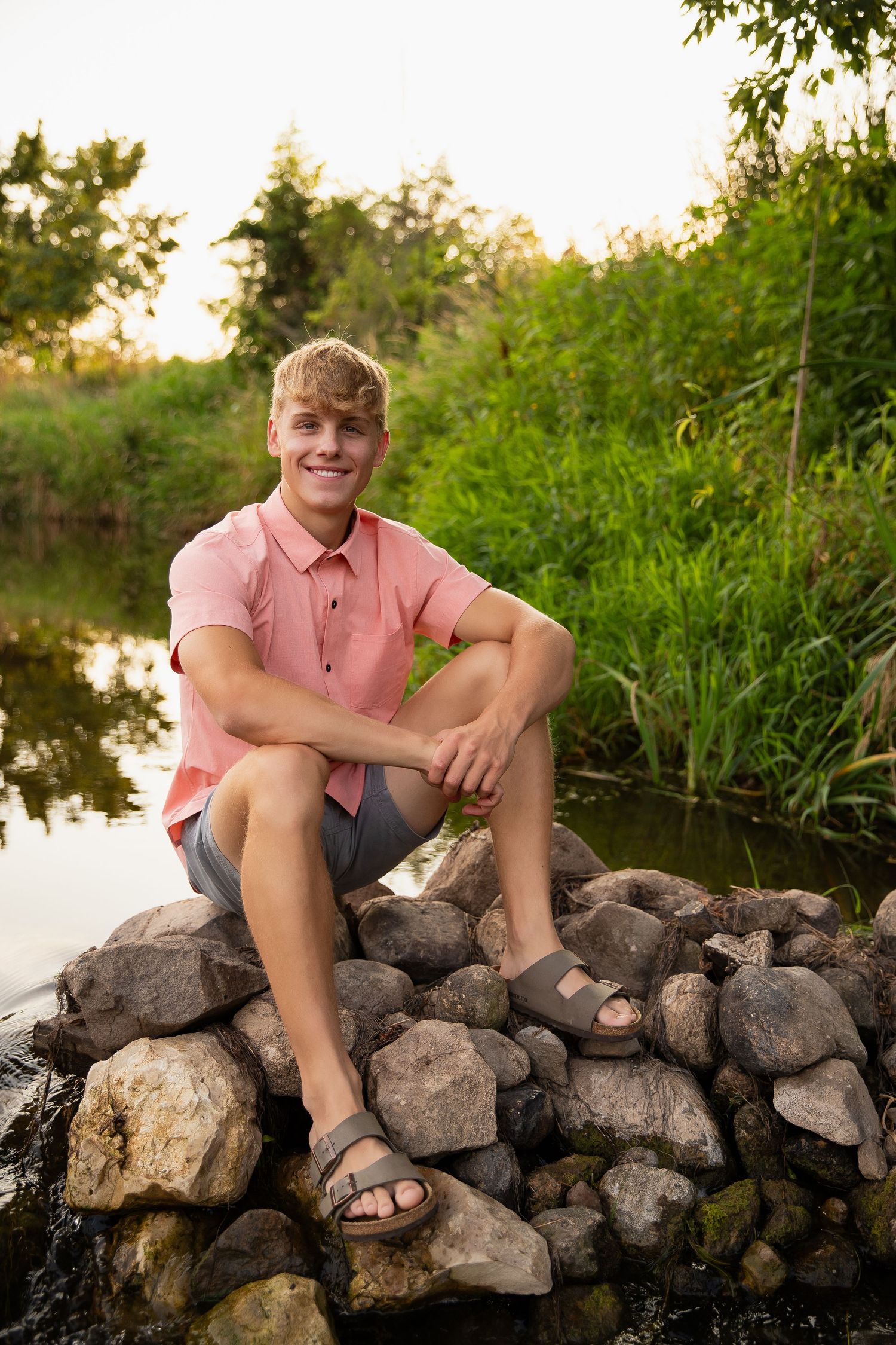 Someone in a pink shirt and sandals sitting on rocks near a tranquil lake at sunset surrounded by lush greenery.