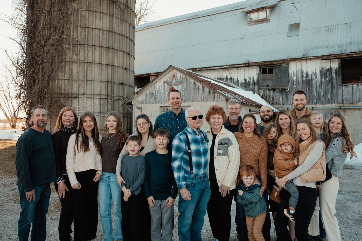 Large family group photo outdoors near a rustic barn and grain silo on a farm during autumn.
