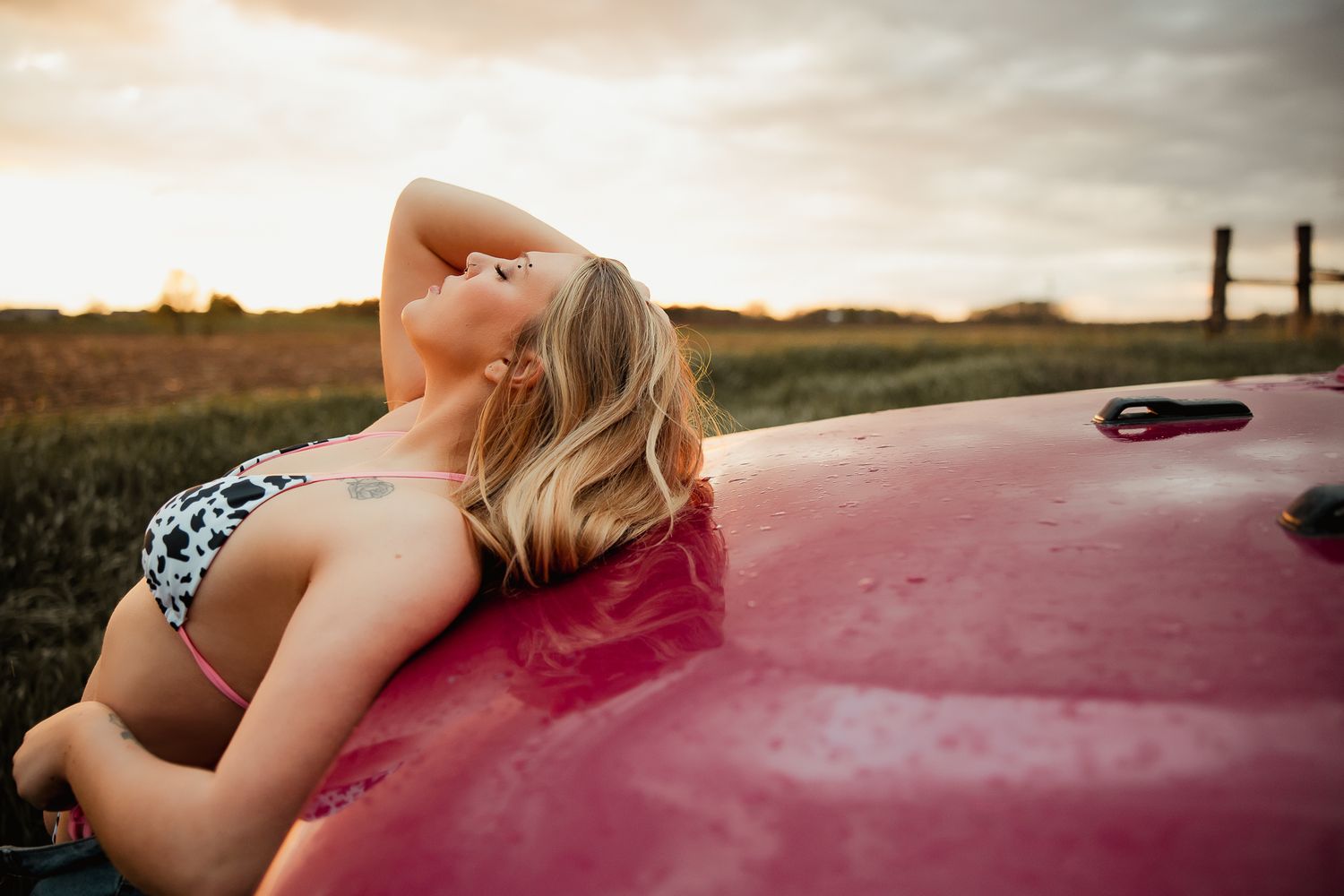 Woman in swimsuit lies back on red car hood during golden sunset in rural field setting.