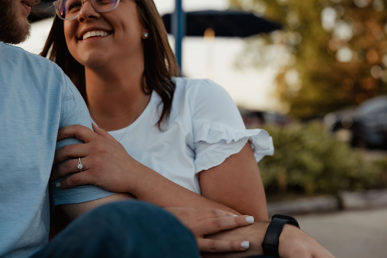Happy couple sitting together outdoors, woman smiling and wearing a ring, man's arm around her shoulder.