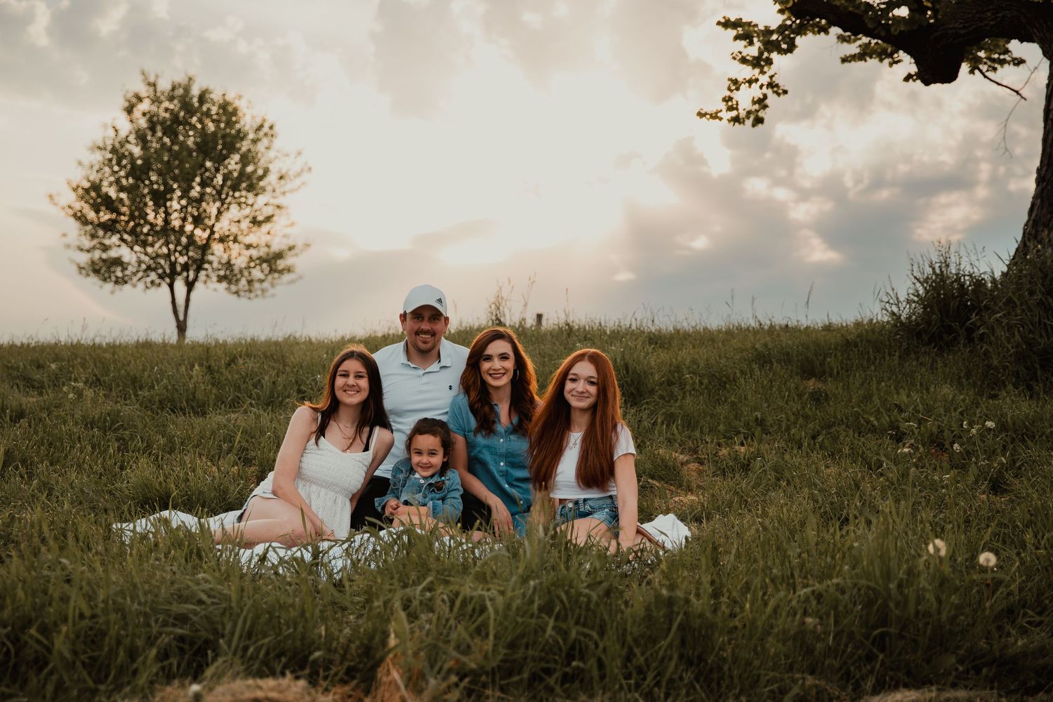 Family of five poses together on a grassy hillside at golden hour, smiling warmly for an outdoor portrait.