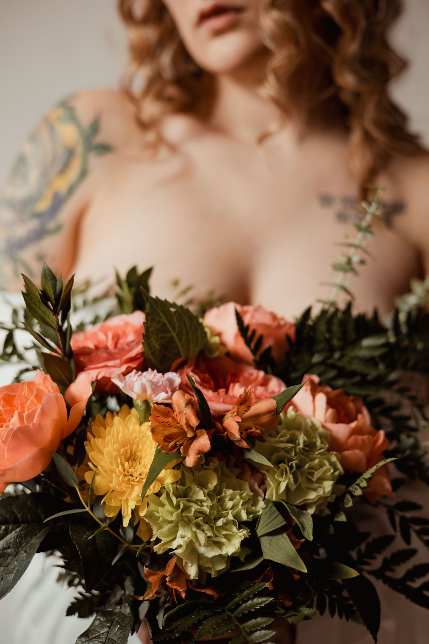 Romantic floral arrangement with coral roses, yellow chrysanthemums and lush greenery against a soft neutral background.