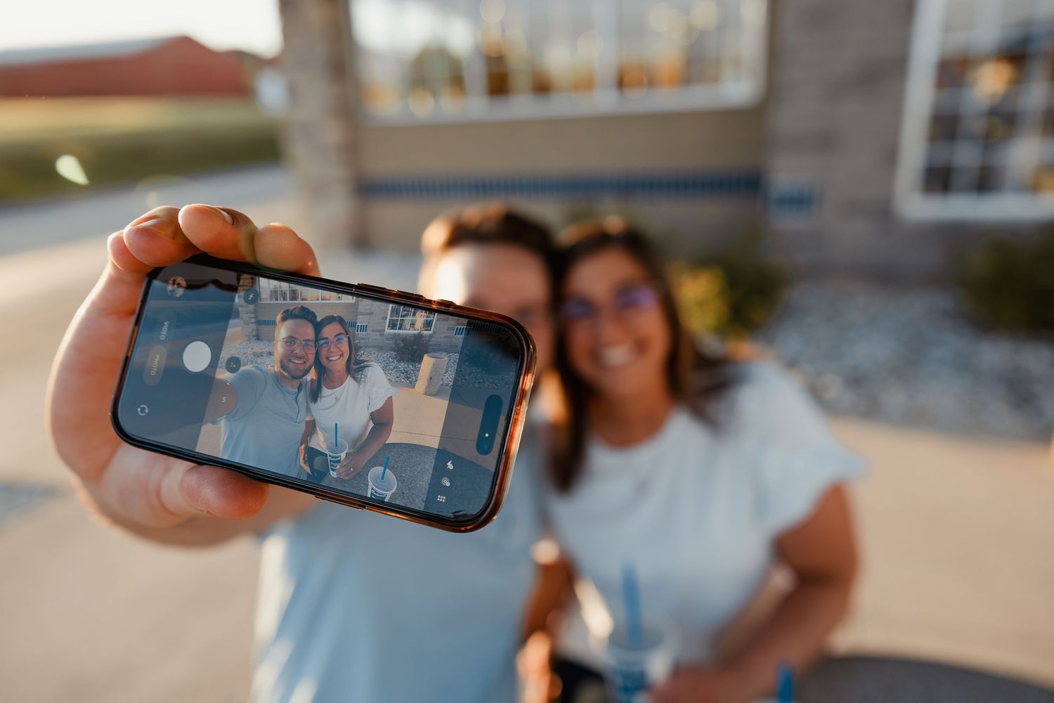 Two people take a selfie outdoors holding up a smartphone showing their photo together.