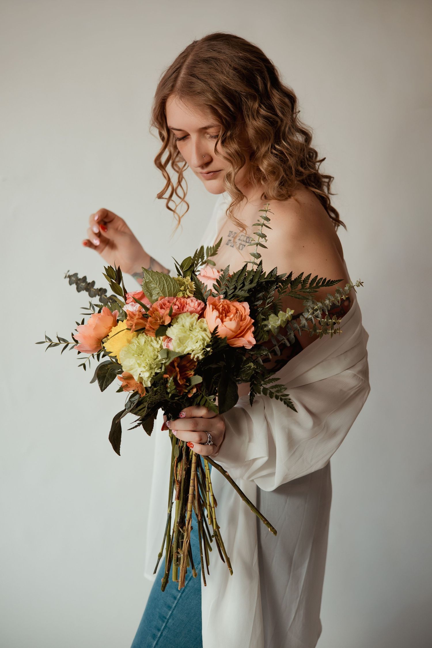 A person in an off-shoulder white top and jeans holds a vibrant bouquet with orange flowers and greenery against a neutral wall.