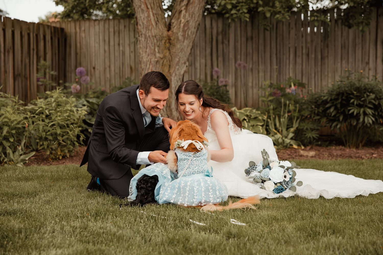 A couple relaxes on green grass near a wooden fence during a formal photoshoot session.