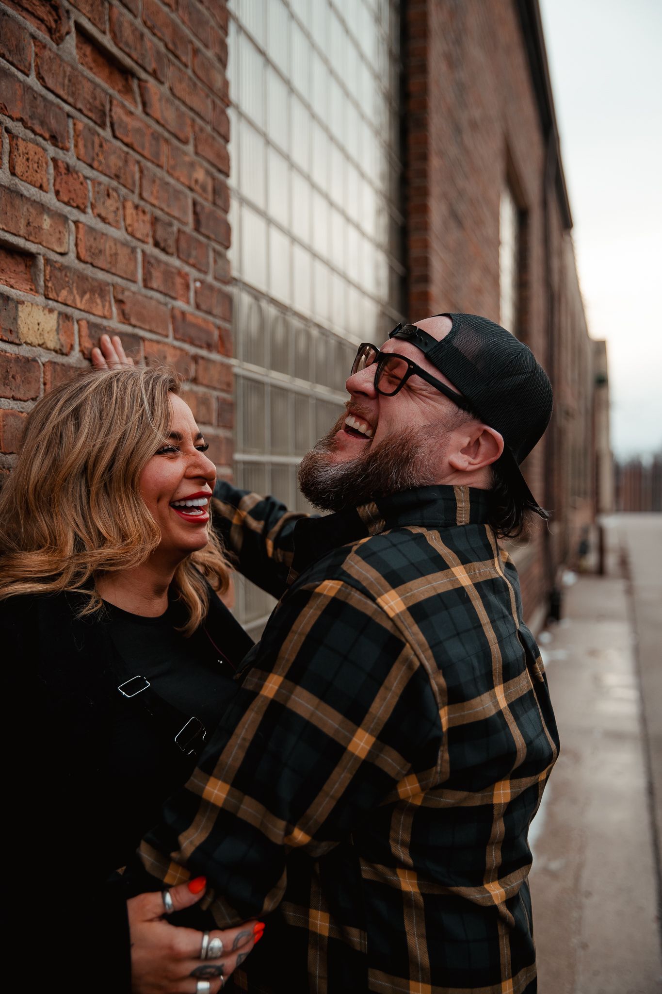 A joyful couple laughs together against a brick wall background in casual autumn attire. engagement couple