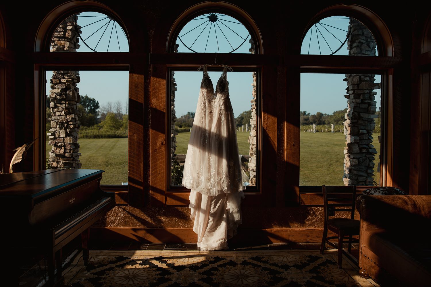 A white wedding dress hangs in an elegant arched window frame overlooking a scenic countryside landscape.