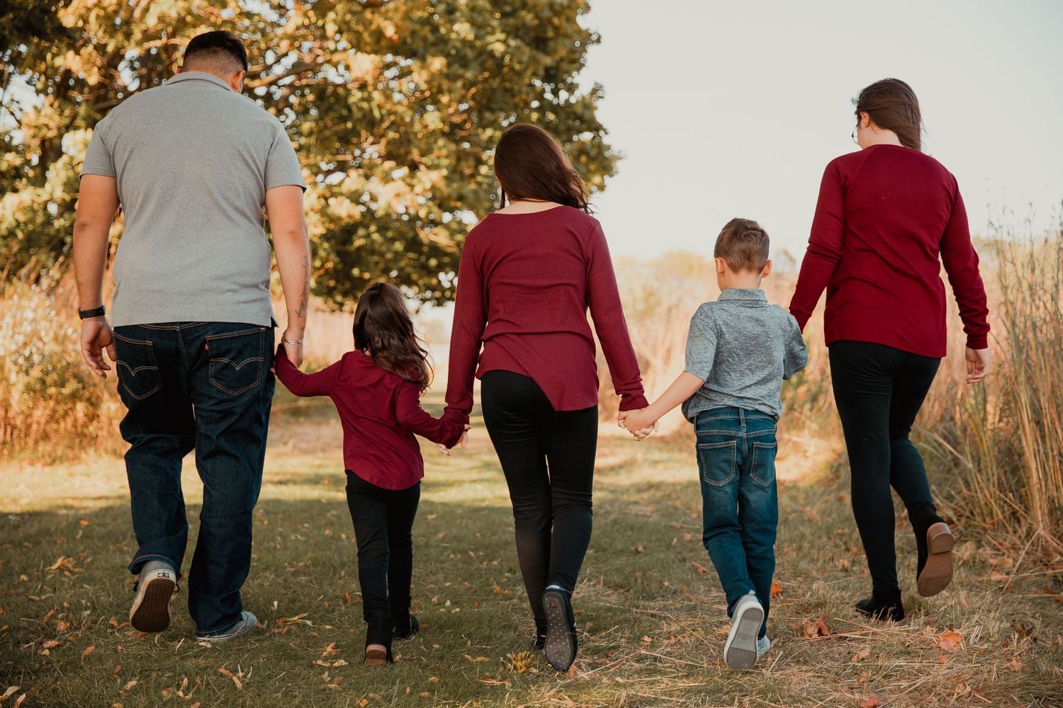Family of five holding hands walking through a scenic autumn field, viewed from behind, wearing coordinated red and blue outfits.