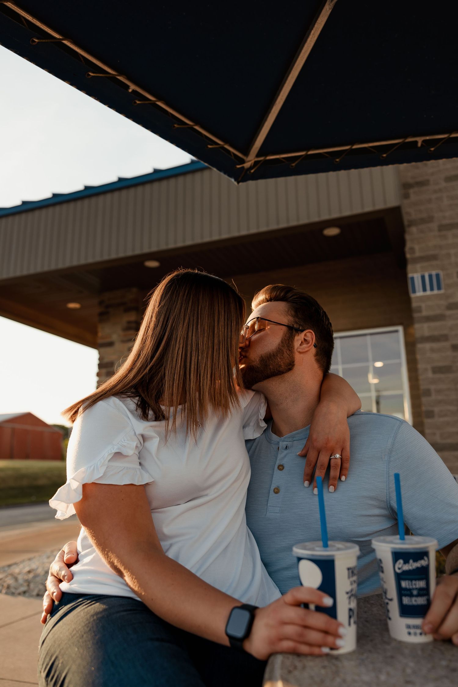 Couple kissing and holding Culvers drinks under a patio umbrella at golden hour.
