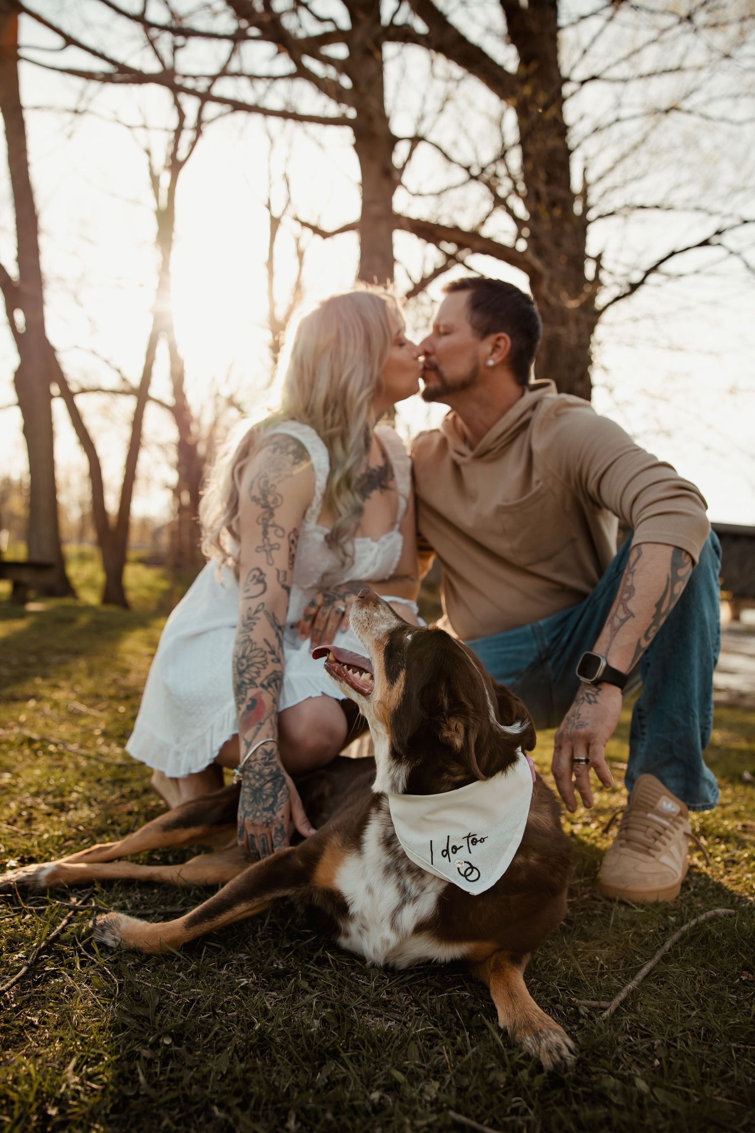 Tattooed couple sharing a kiss at golden hour in a park, with their dog wearing an I do too engagement bandana.