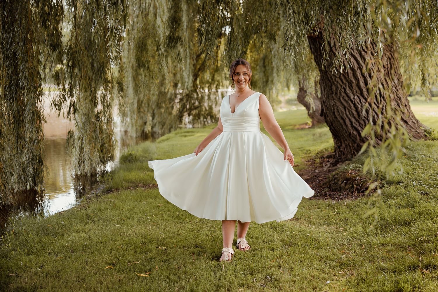 Person in a white vintage-style dress twirling in a garden setting with willow trees and a pond.