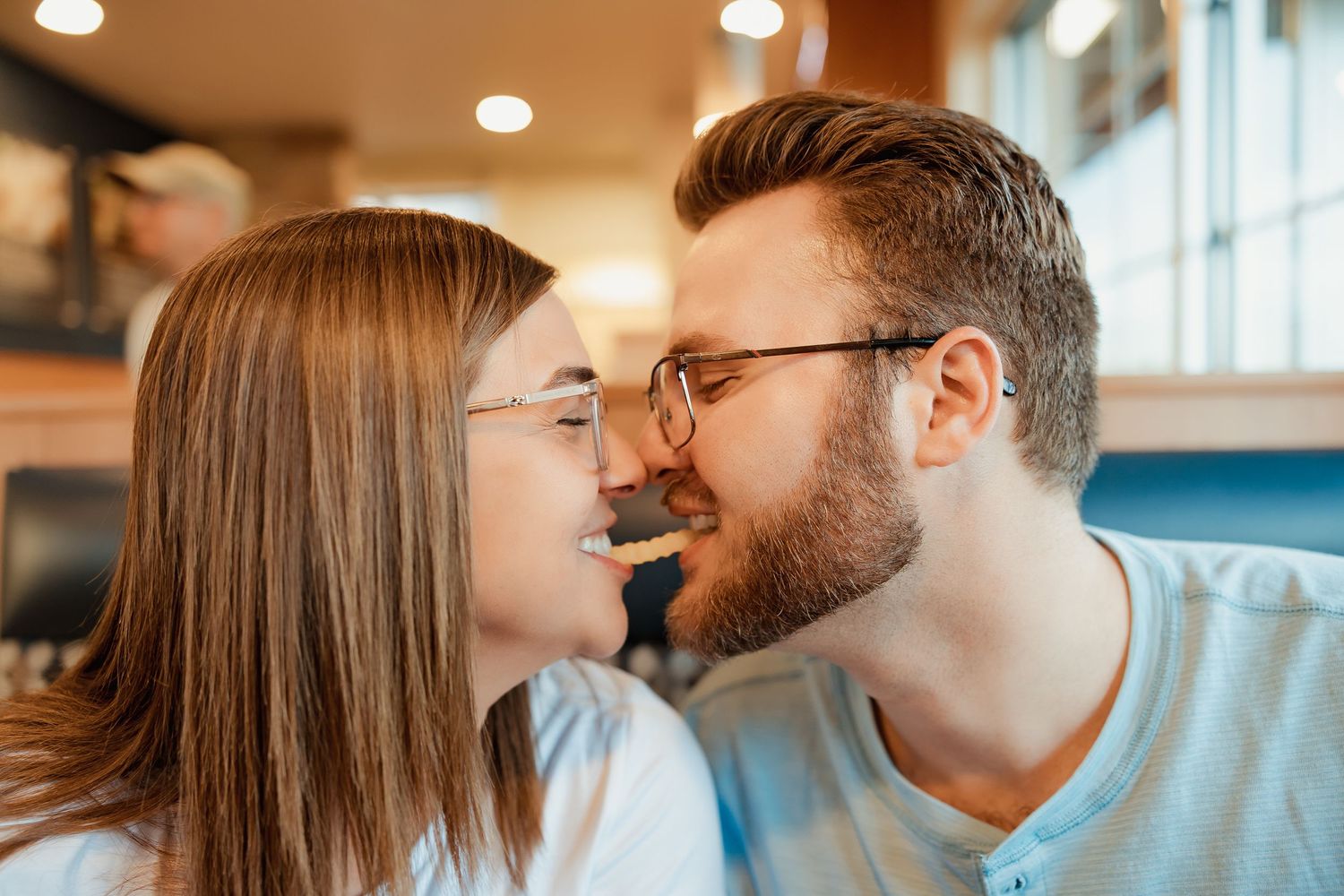 A couple sharing a romantic moment, nearly kissing while wearing glasses at a casual restaurant.