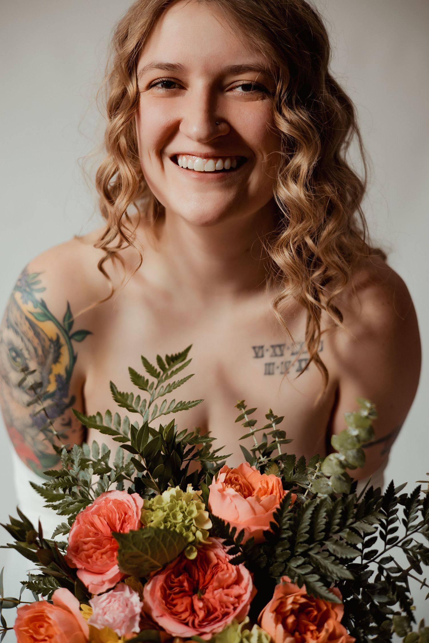 Portrait with curly hair and colorful floral bouquet featuring coral roses and ferns against neutral background.