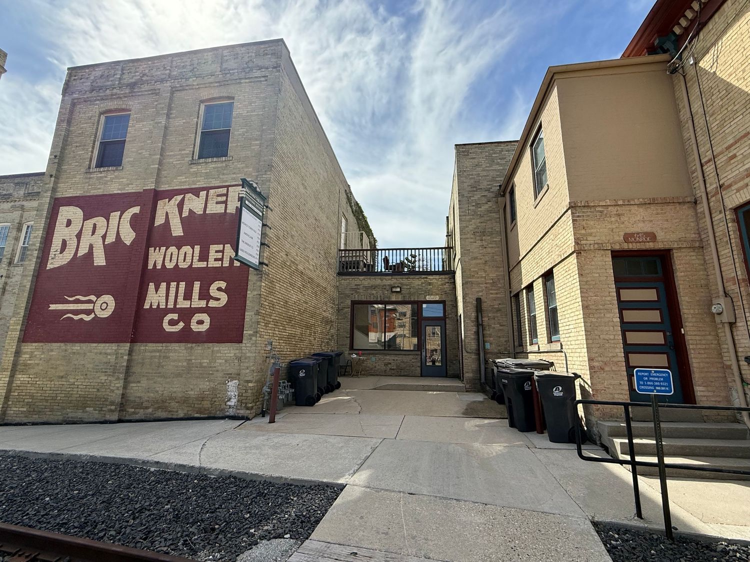 Historic brick building with faded Brick Woolen Mills Co signage and alleyway between commercial structures in urban setting.