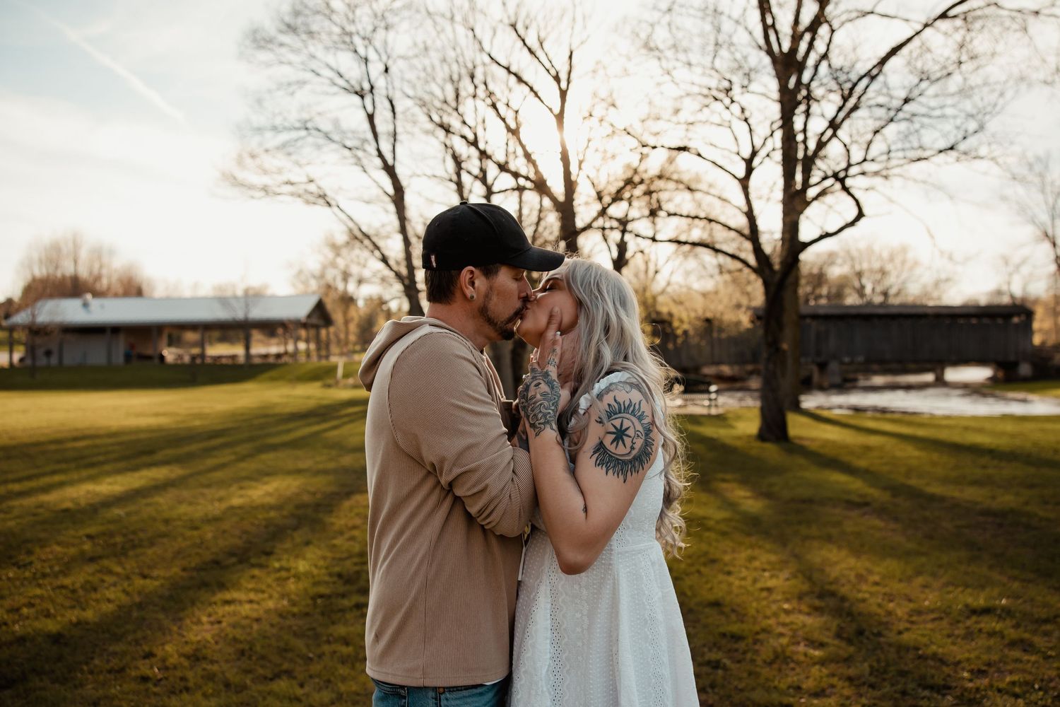 Couple sharing a romantic forehead kiss outdoors at golden hour in a lush green park with bare trees.
