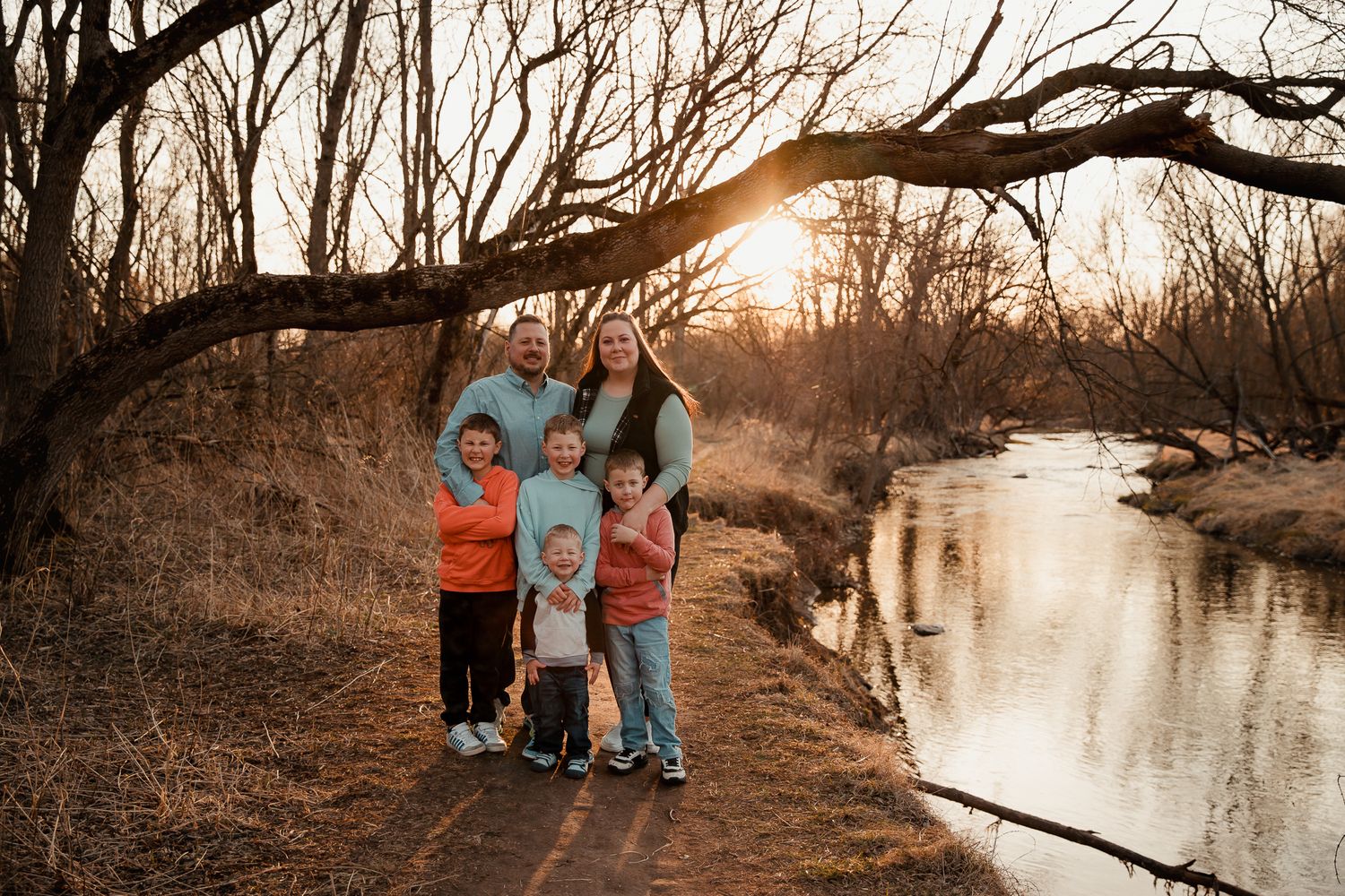 Family of five posing by a creek at golden hour sunset surrounded by bare winter trees.