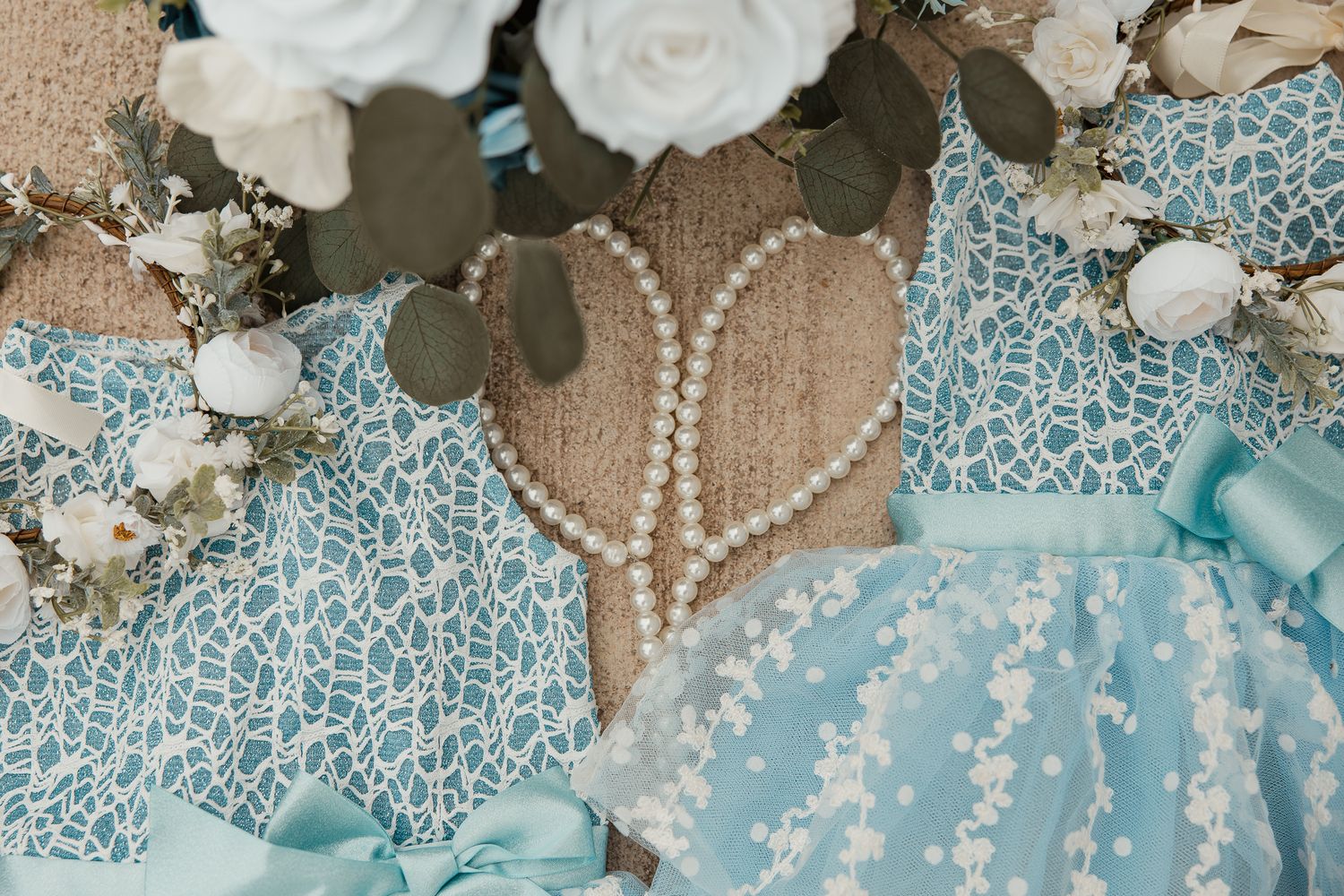 Elegant light blue lace dresses with pearl details and white roses arranged on a champagne-colored background.