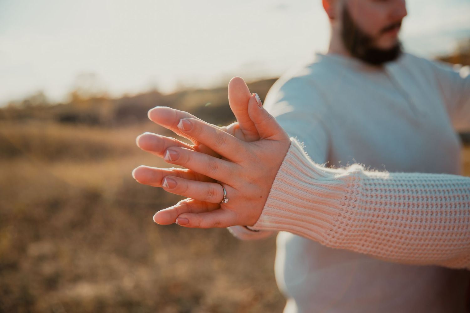 Close-up of hands wearing a warm cream sweater against a sunset backdrop. couples engagement