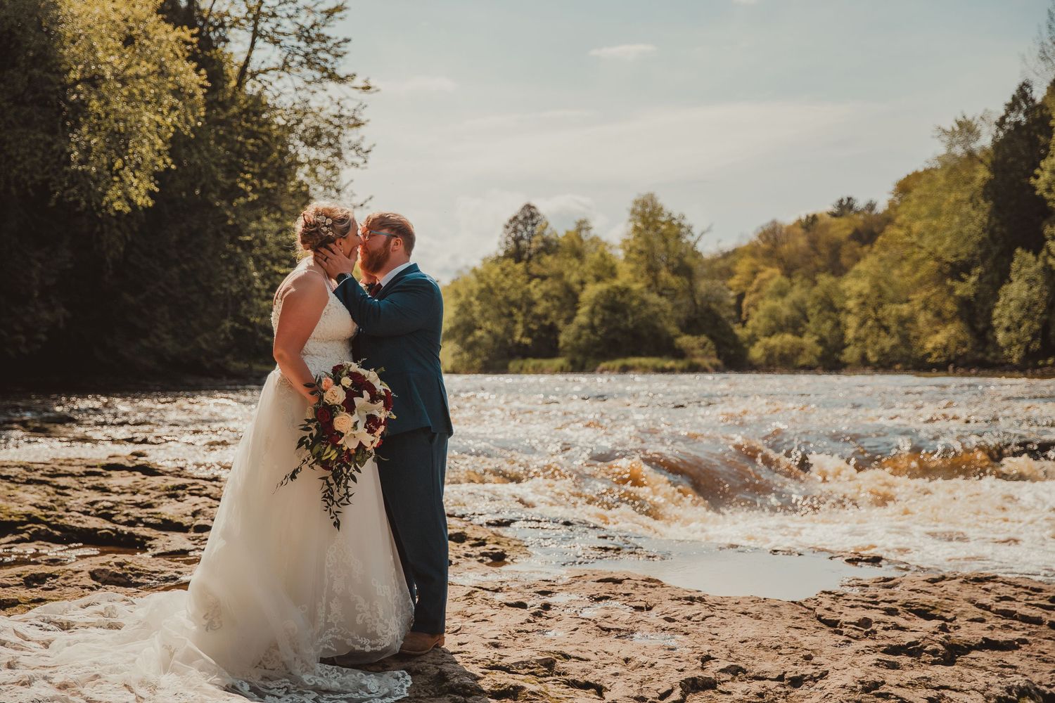 A romantic wedding photo of a couple embracing by a scenic riverside at sunset. wedding