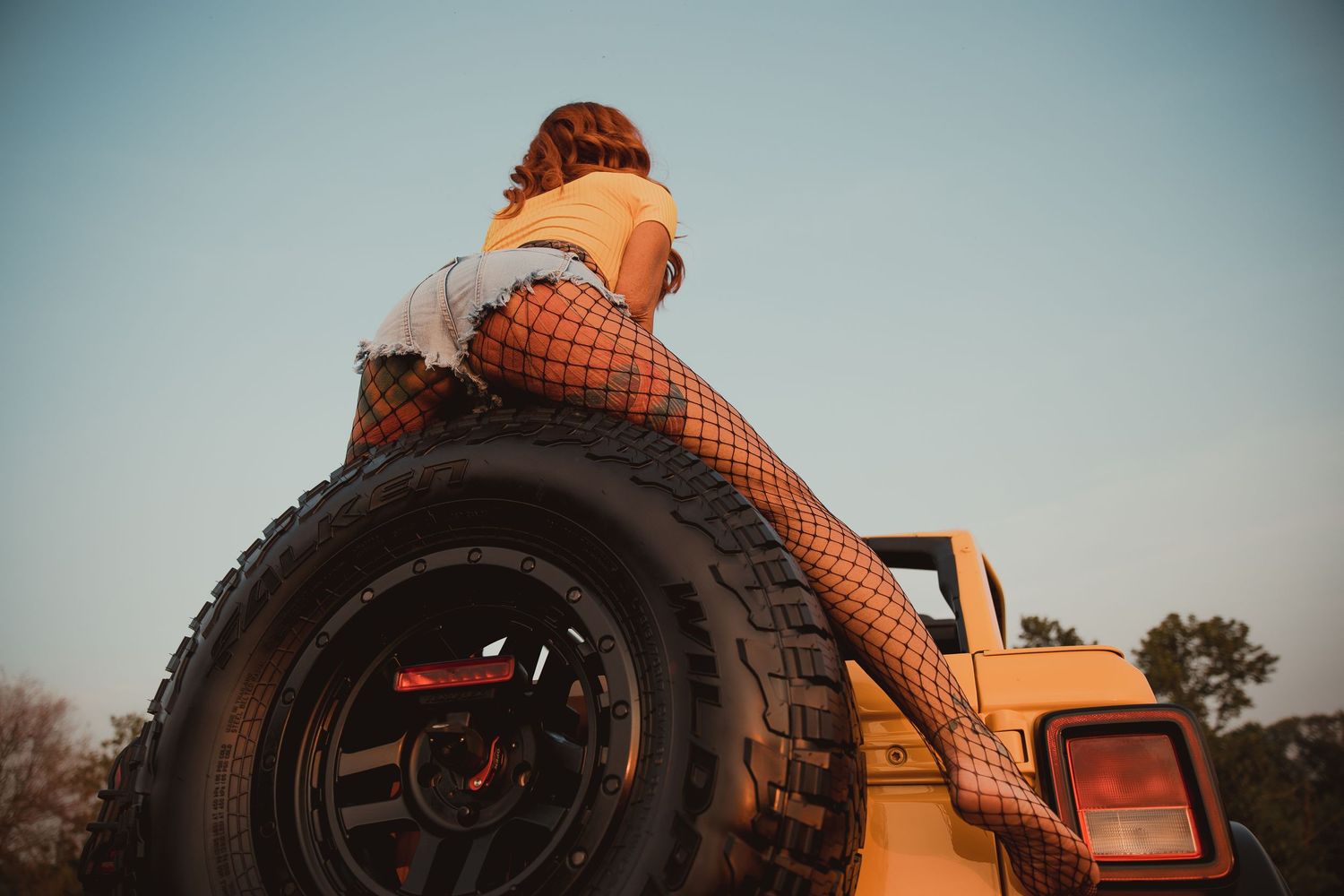 Person in plaid outfit sits atop a large offroad tire on a vintage tractor at sunset.