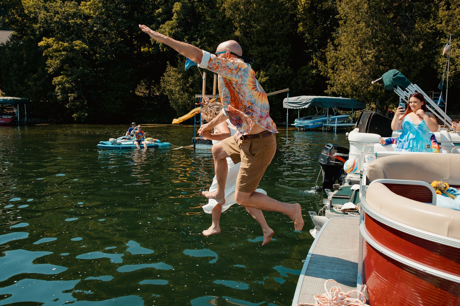 People jumping off a dock into a lake surrounded by trees and boats on a sunny summer day.