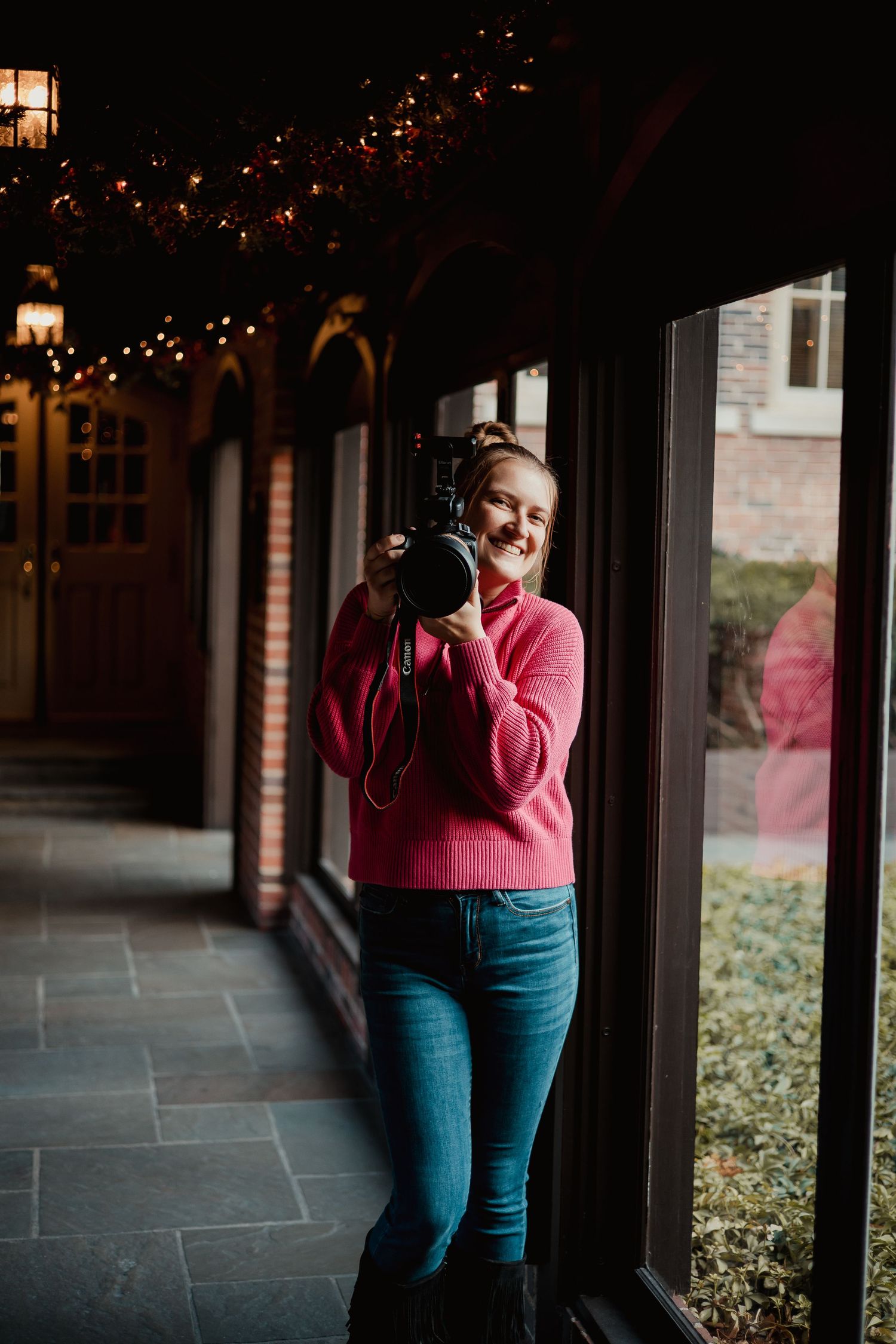 A person in a pink sweater and blue jeans stands in a covered walkway with string lights overhead.