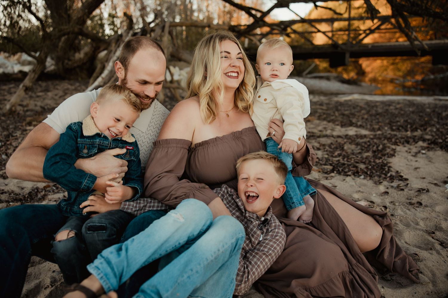 Happy family of five sitting outdoors on sandy ground, laughing together during a warm golden hour photo session.