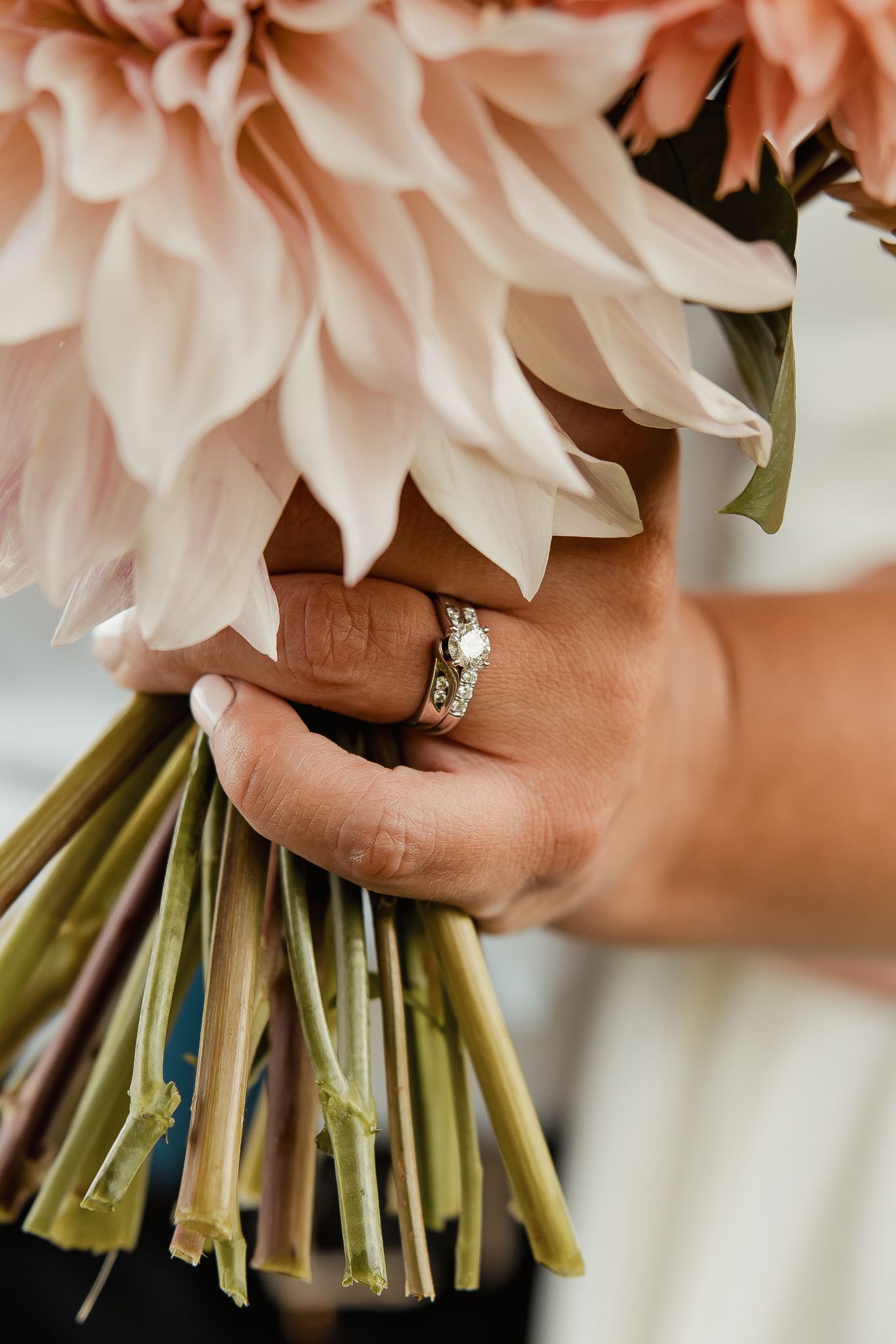 Close-up of hands holding a blush pink flower bouquet with a diamond wedding ring visible.