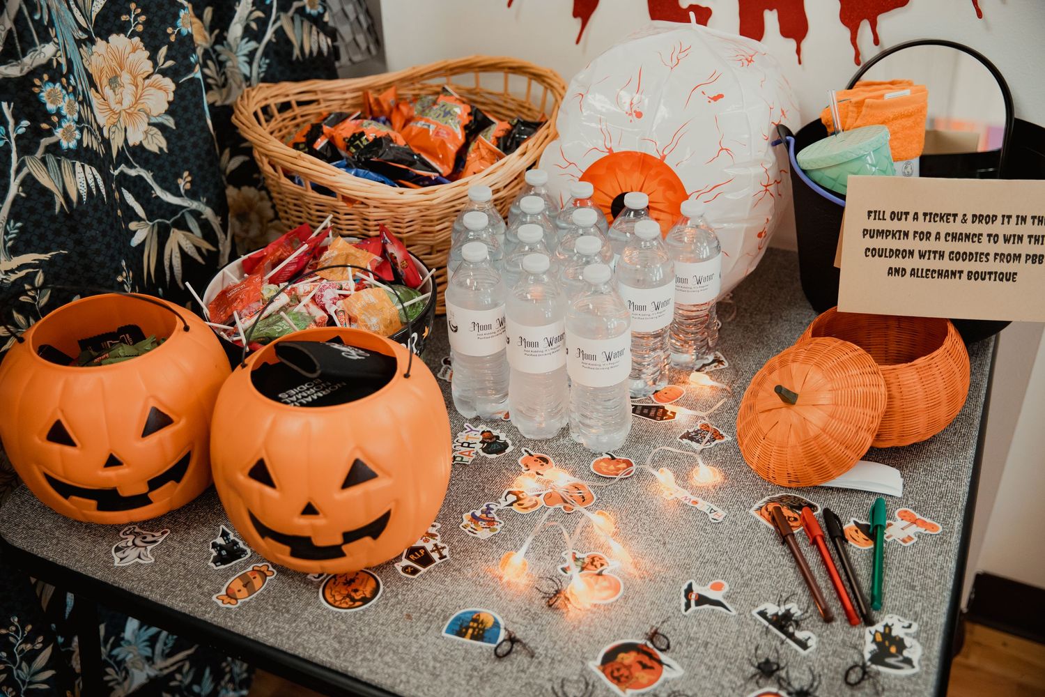 Halloween party table with plastic pumpkin buckets, candy, water bottles, and spooky decorations on display.