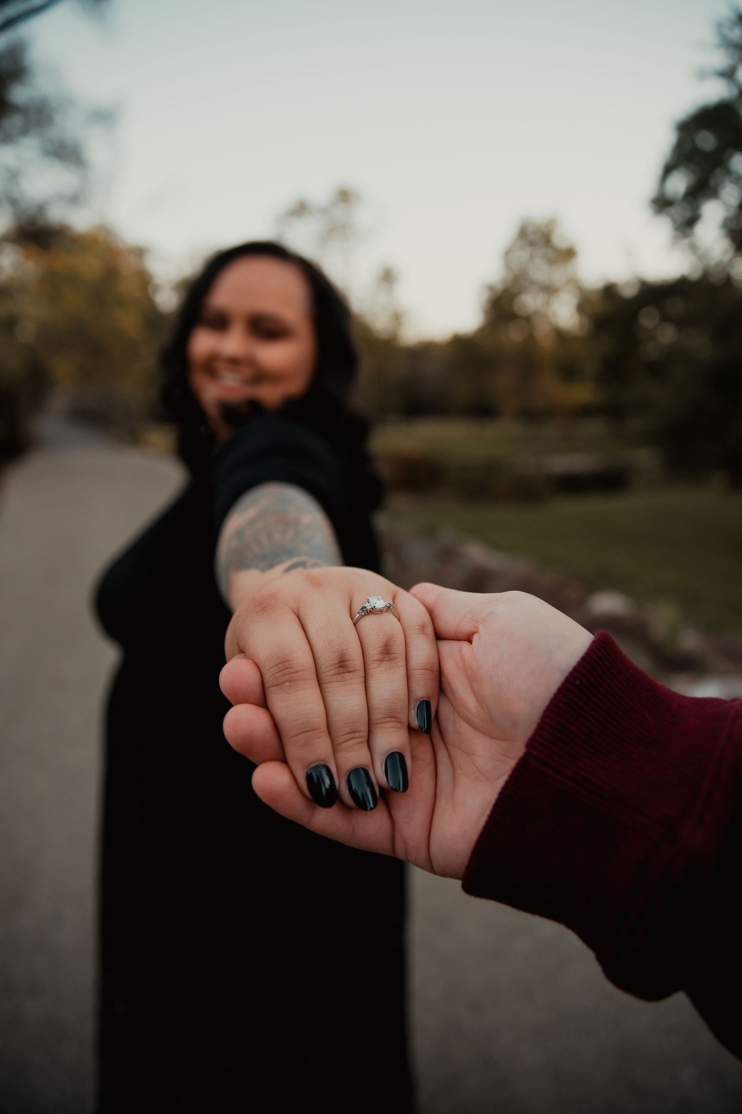 Close-up of clasped hands showing an engagement ring with dark nail polish against a blurred autumn background. couples engagement