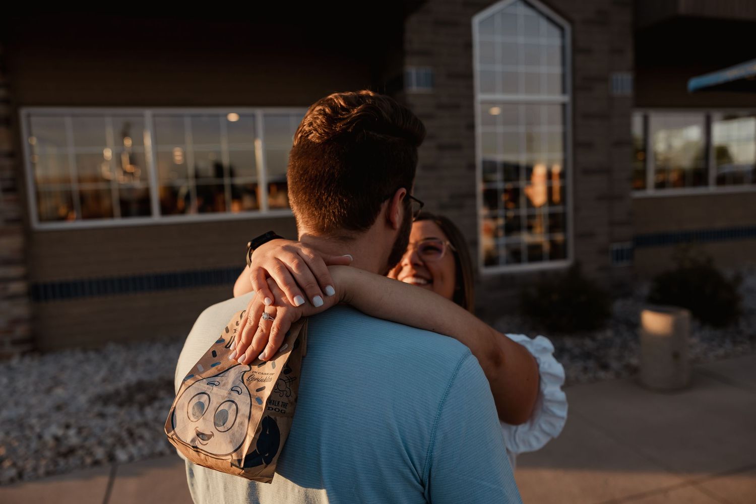 Couple embracing outdoors at sunset near a building, woman wearing white top hugging man with backpack.