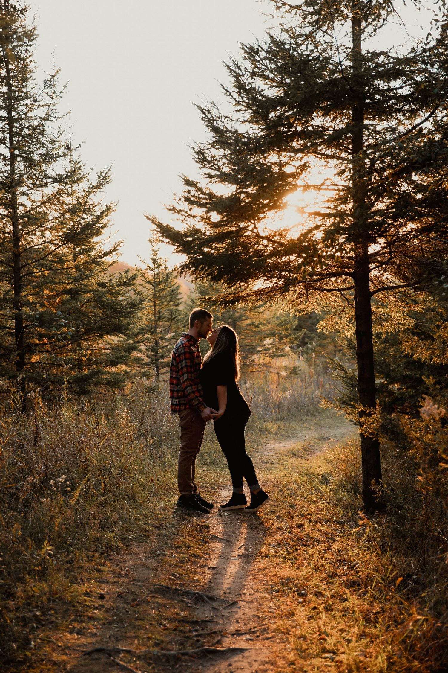 Two silhouettes embrace on a forested path at sunset with golden light filtering through evergreen trees.