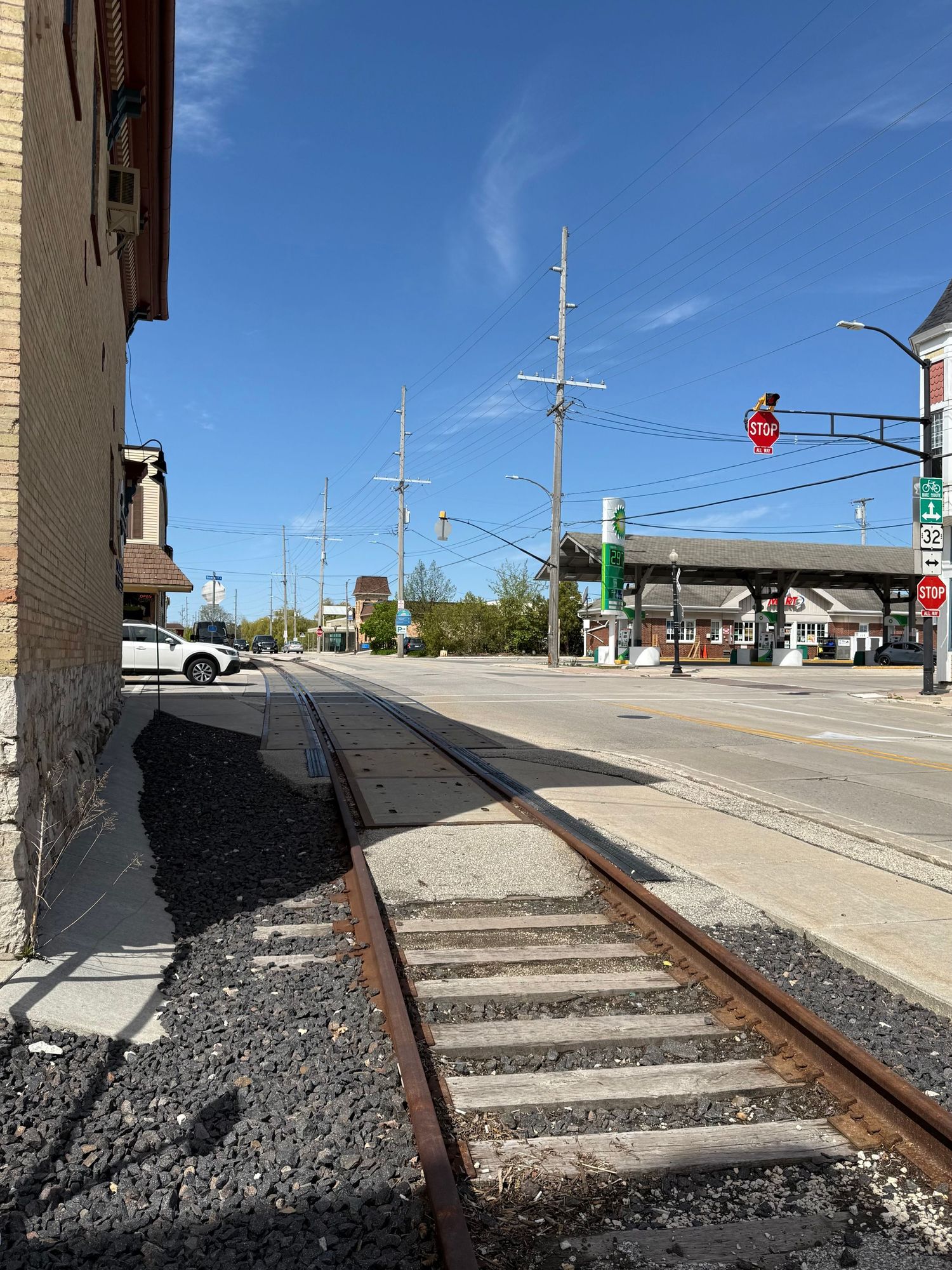 Railroad tracks run along a city street with traffic lights and utility poles under a bright blue sky.