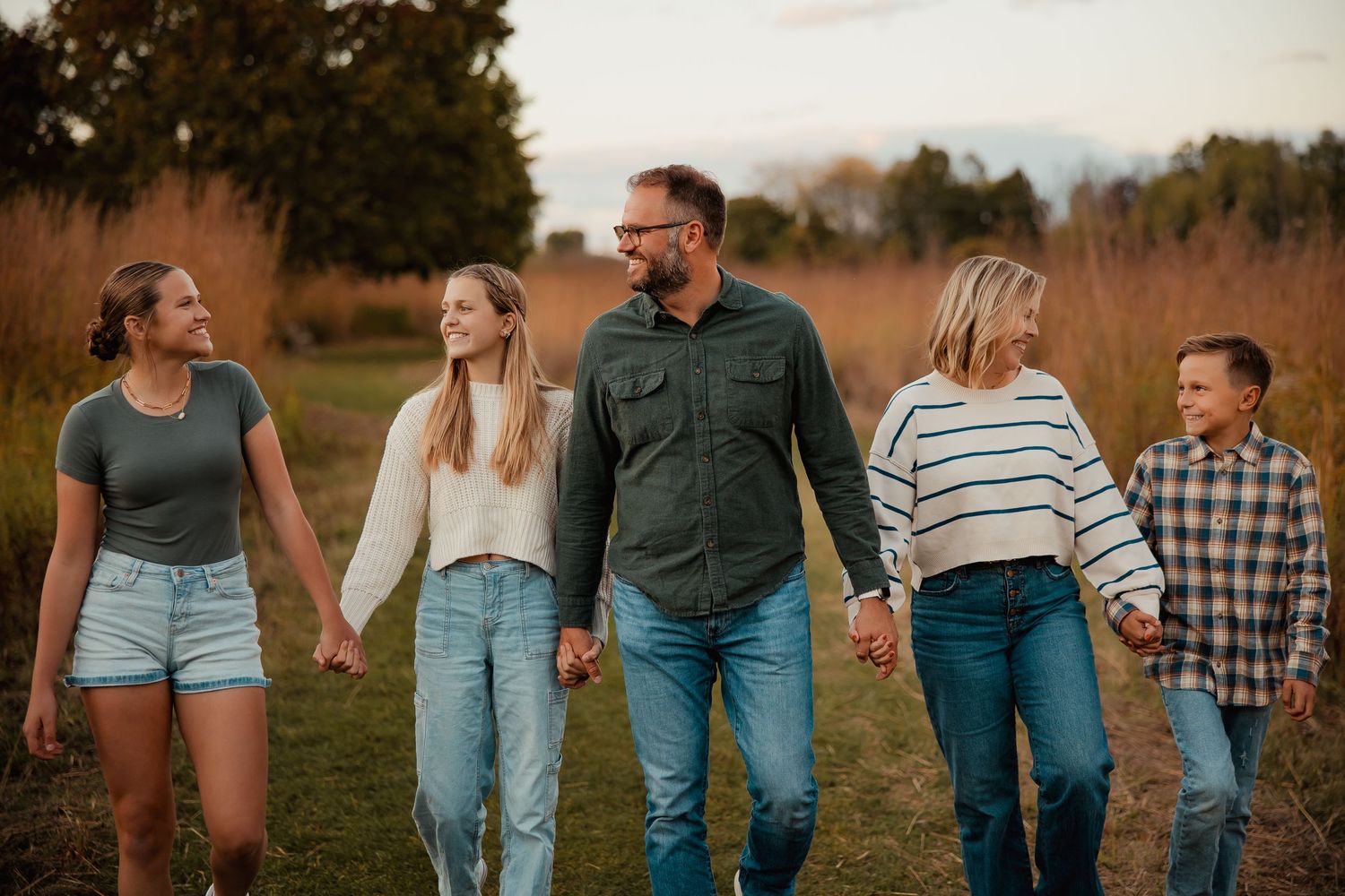 Family of five walking hand-in-hand through a golden autumn field, smiling and enjoying quality time together outdoors.