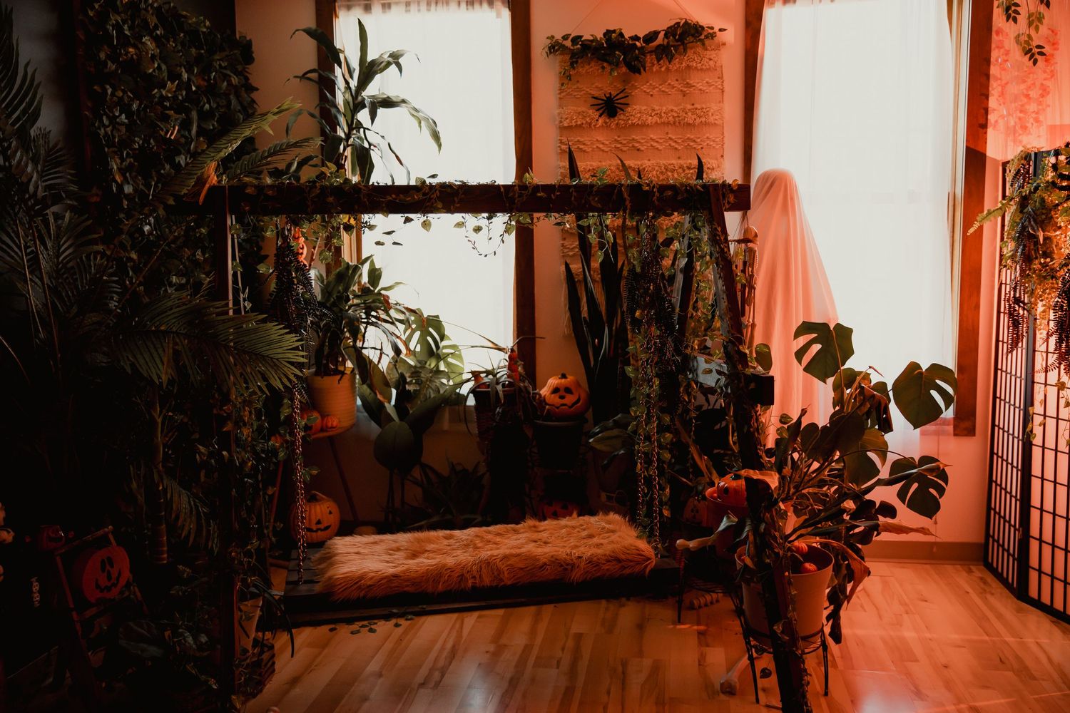 Indoor swing seat surrounded by potted plants and houseplants in a warm orange-lit room with wooden flooring.